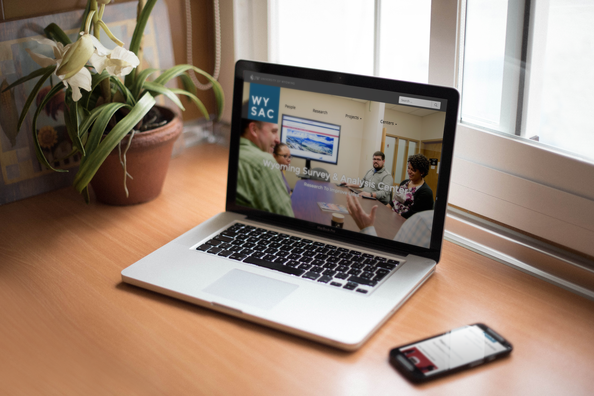 A laptop and smartphone sitting on a desk next to a plant. The redesigned WYSAC website is on the screen.
