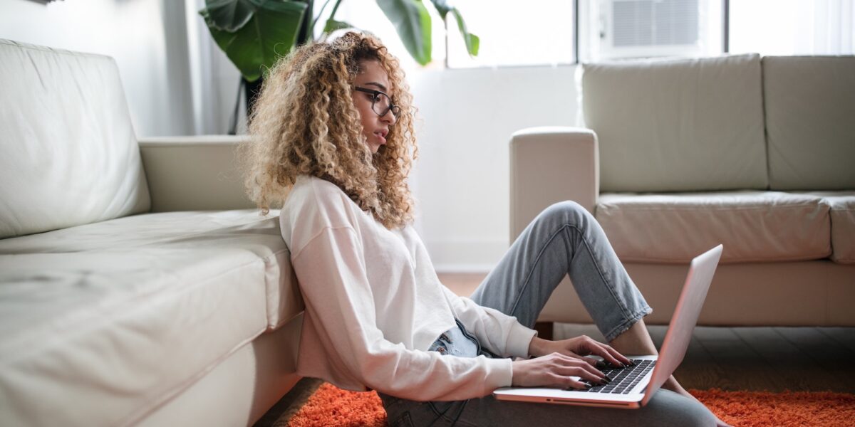 Young woman with curly hair and glasses working on a laptop while sitting on the floor, leaning her back on the couch