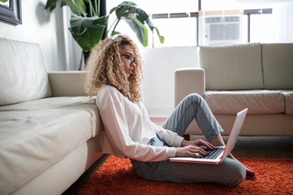 Young woman with curly hair and glasses working on a laptop while sitting on the floor, leaning her back on the couch