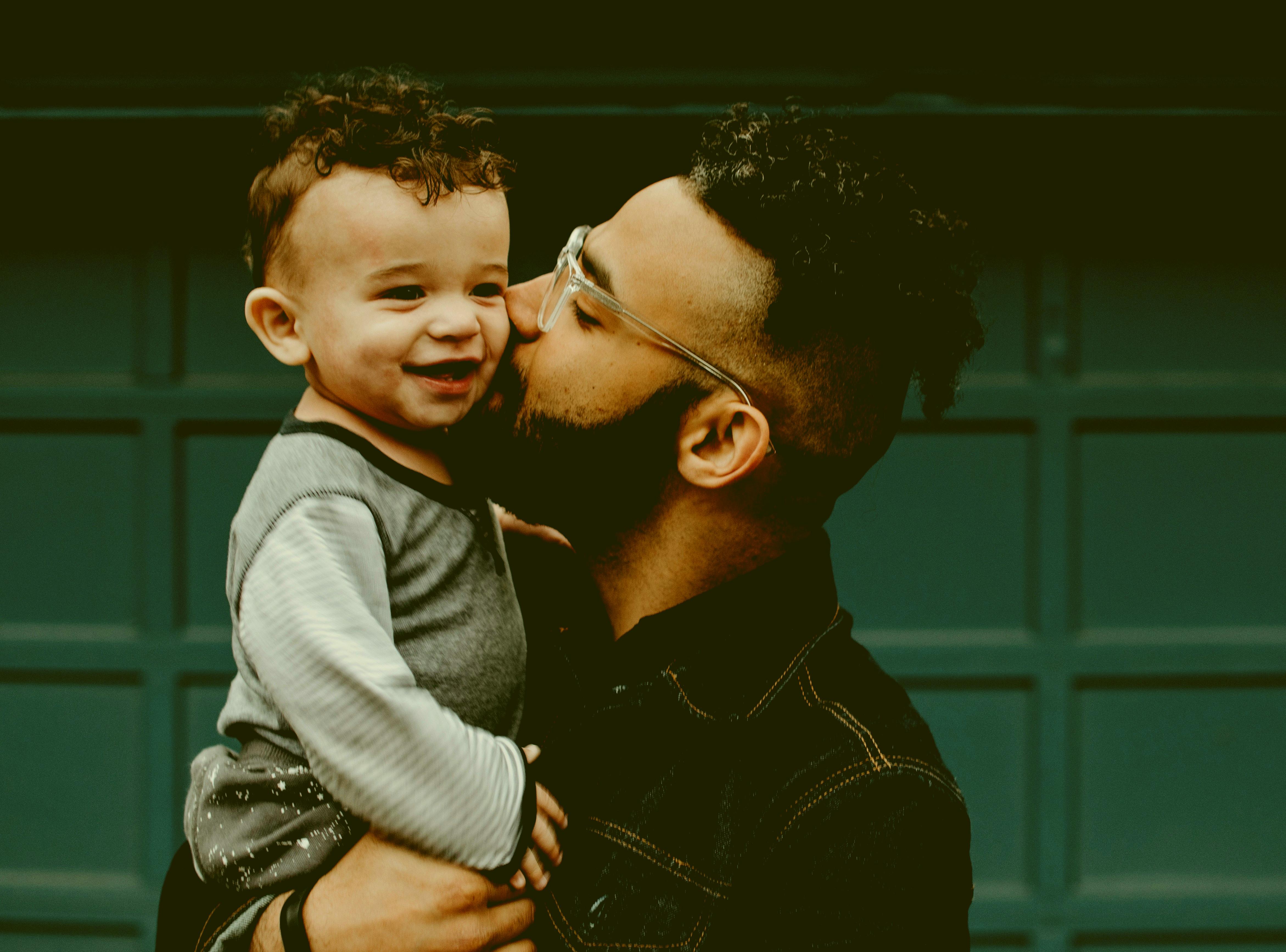 A dad holding a his little, happy baby, giving him a kiss on the check.